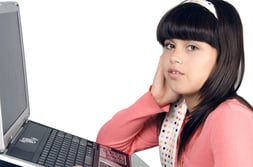 Pretty brunette schoolgirl in pink looks at you, notebook computer in front of her, isolated on white