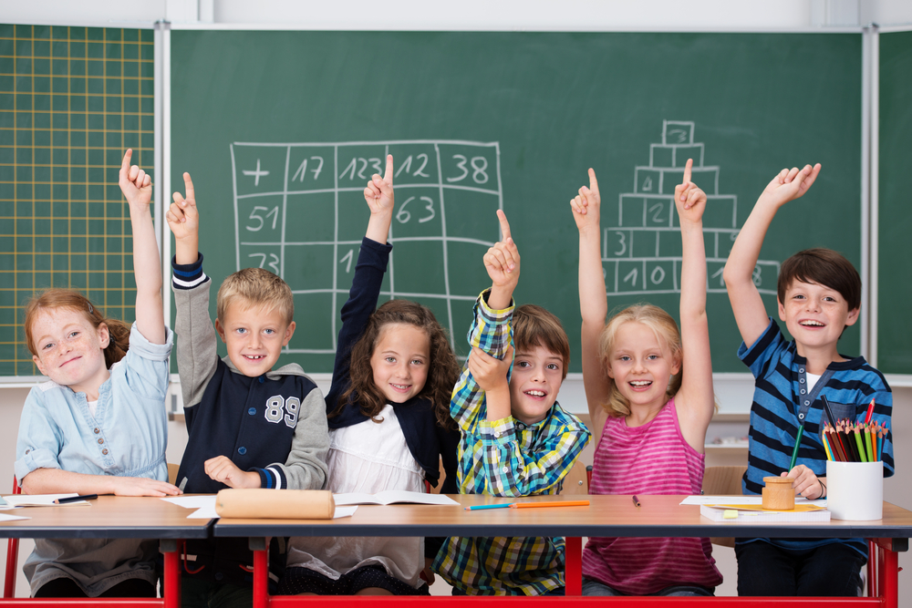 Enthusiastic group of young kids in class sitting in a row at their desk raising their hands in the air to show the know the answer to a question
